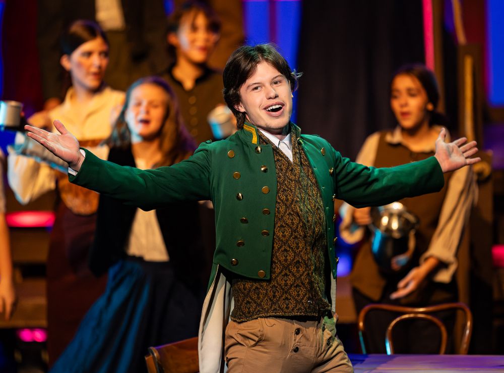Actor in green coat performing energetically with ensemble cast in background.