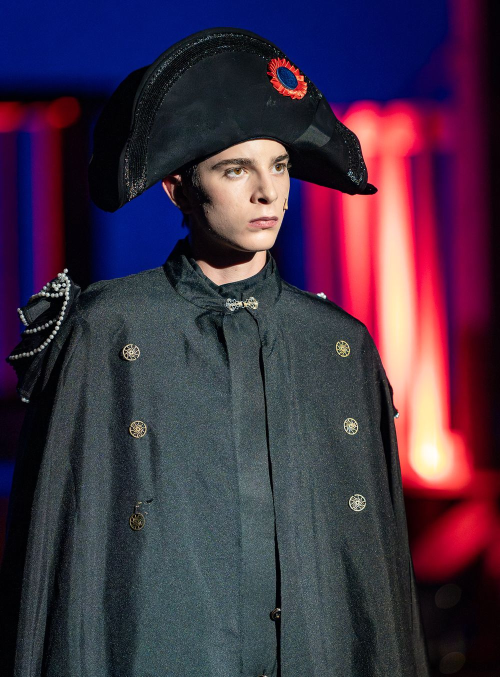 Actor in military-style costume wearing black bicorn hat against red stage lighting.
