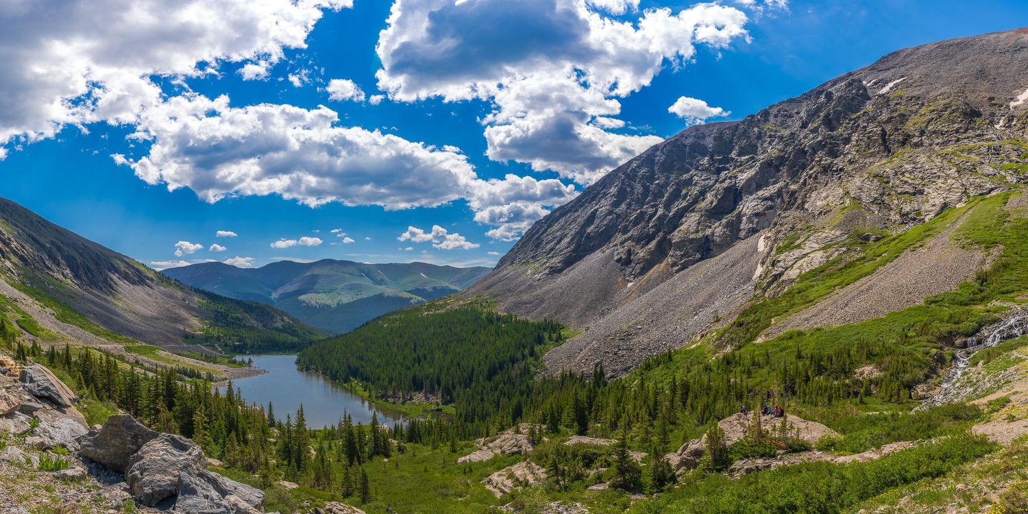 A scenic mountain panorama overlooking a turquoise alpine lake surrounded by evergreen forests and rocky peaks under puffy clouds.