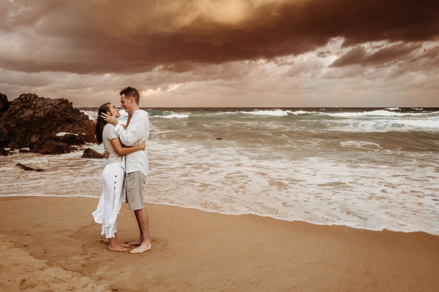A couple embraces on a beach at sunset with dramatic storm clouds overhead.