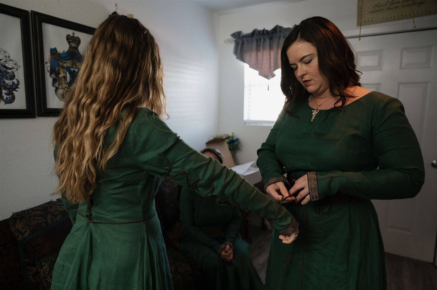 Two figures in matching emerald green medieval-style dresses adjusting their outfits.