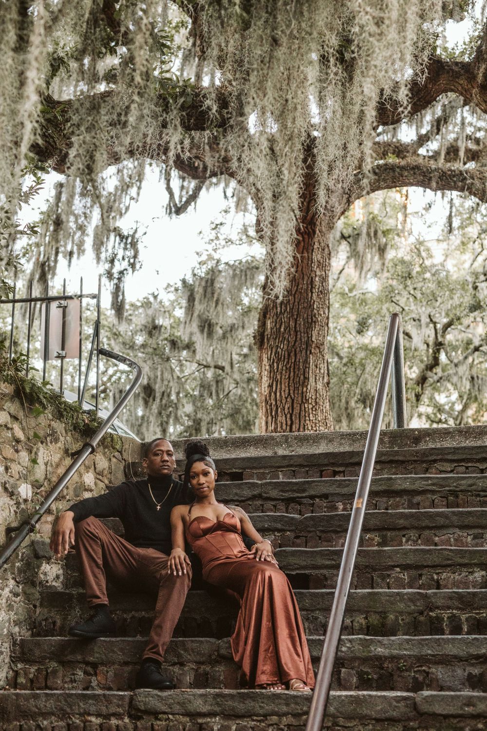 Person in burgundy dress poses on stone steps under Spanish moss draped trees.