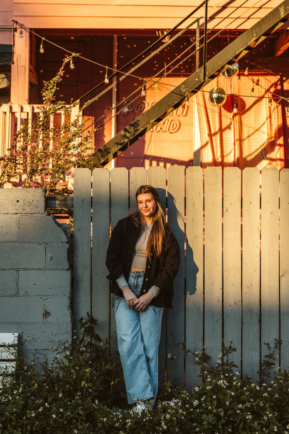 Couple poses against white fence during sunset engagement photoshoot.