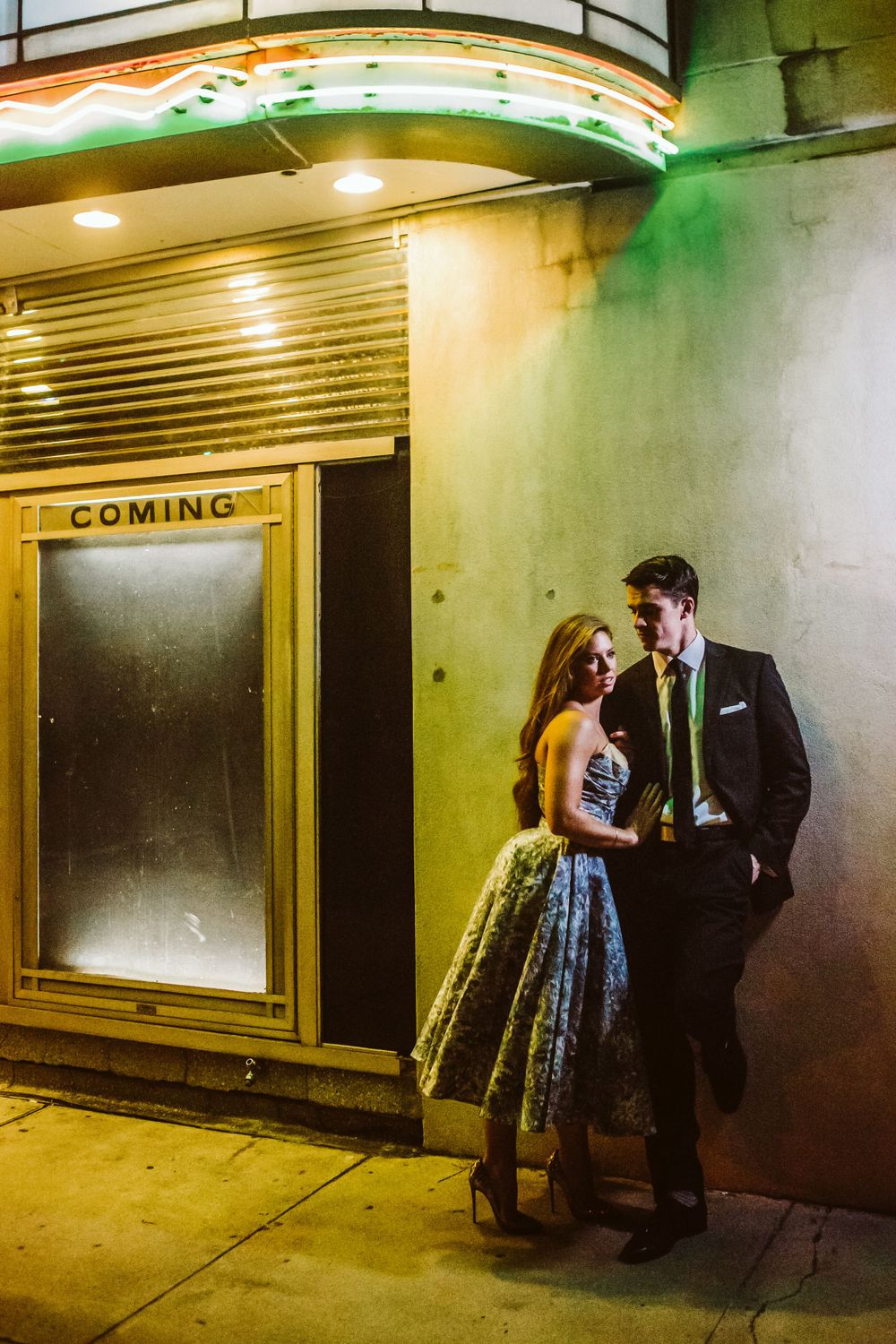 A couple embraces in a dimly lit industrial hallway with green lighting.