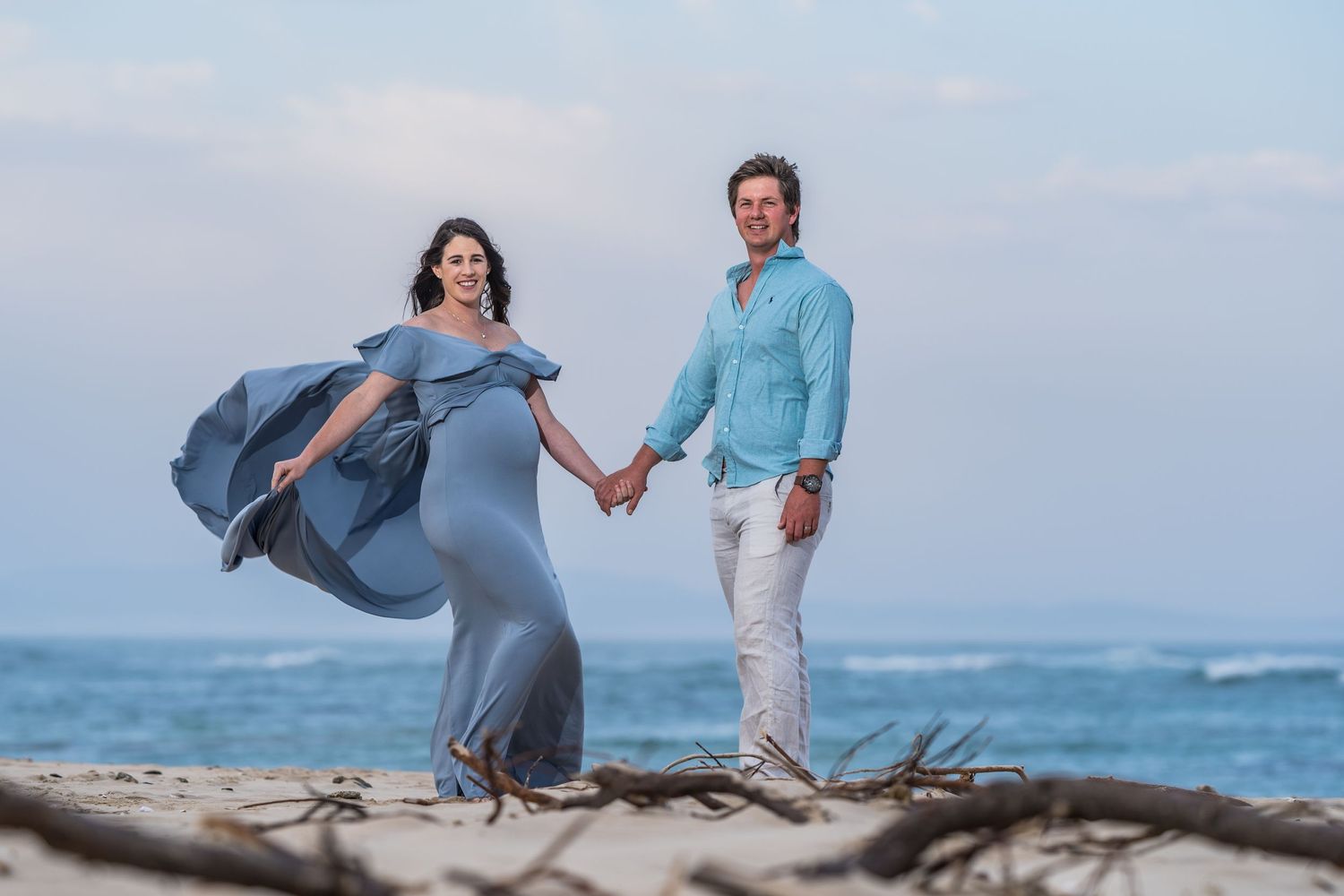 Pregnant woman in elegant blue maternity gown walks hand in hand with partner on a scenic beach.