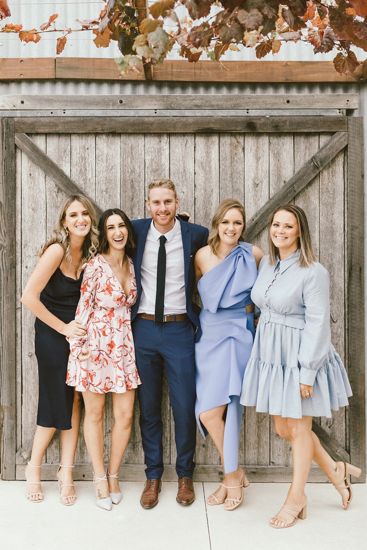 Wedding party sitting together against wooden fence in coordinated blue and floral outfits.
