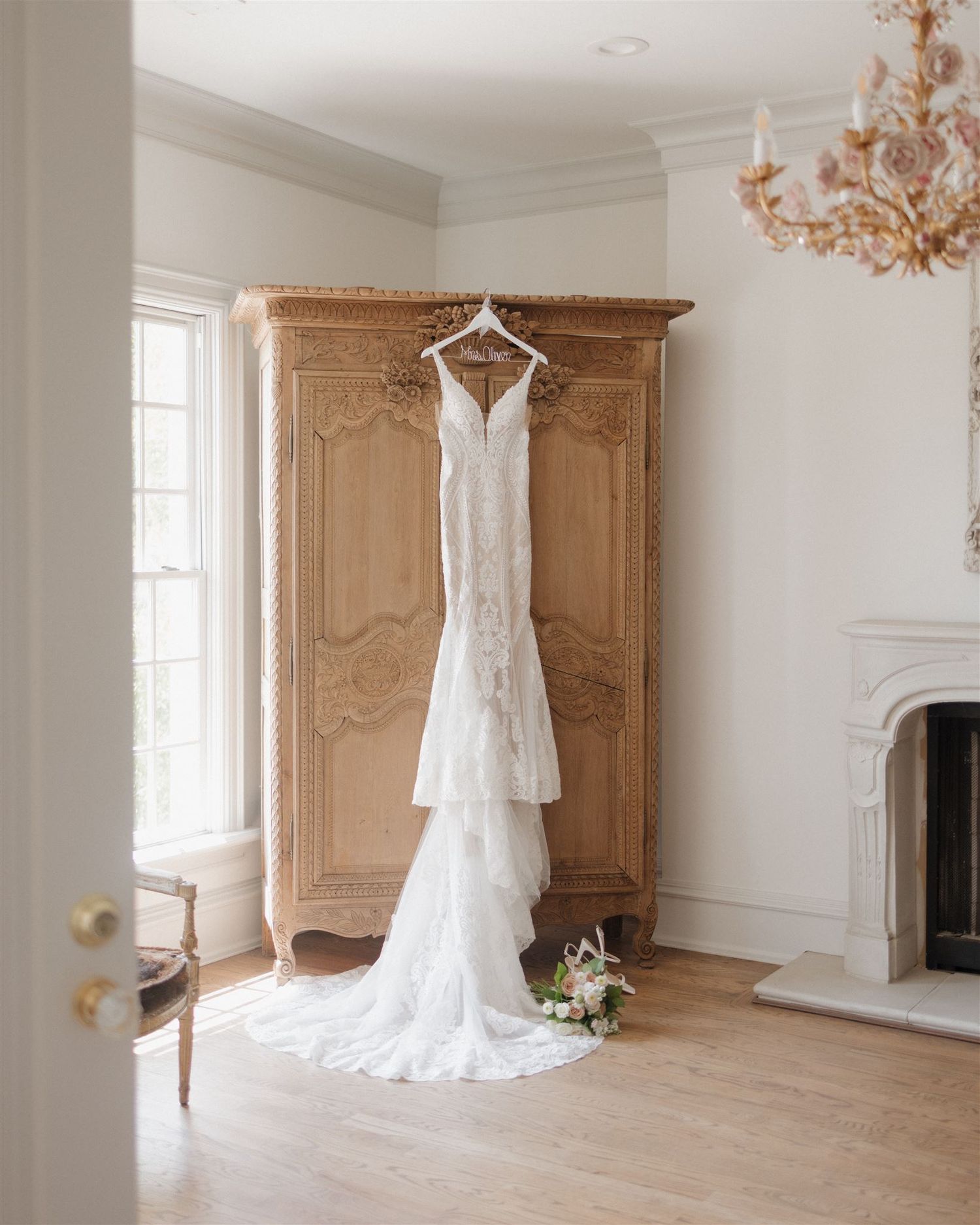 A lace wedding dress hangs elegantly on a wooden armoire next to a fireplace in a bright room.