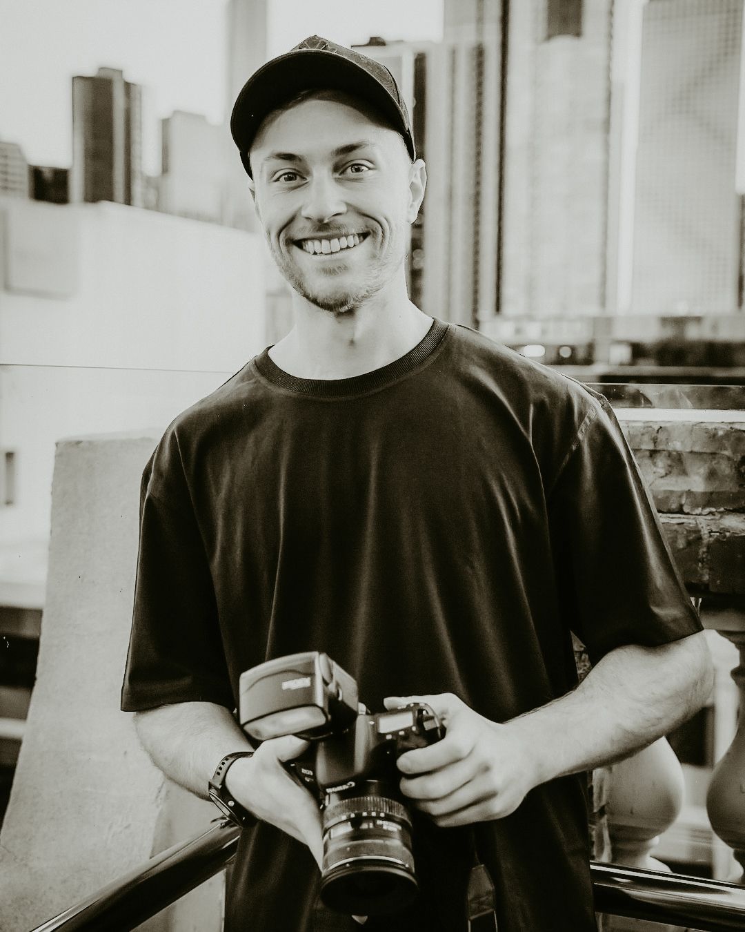 Black and white photo of a photographer wearing a baseball cap and holding a camera while smiling.