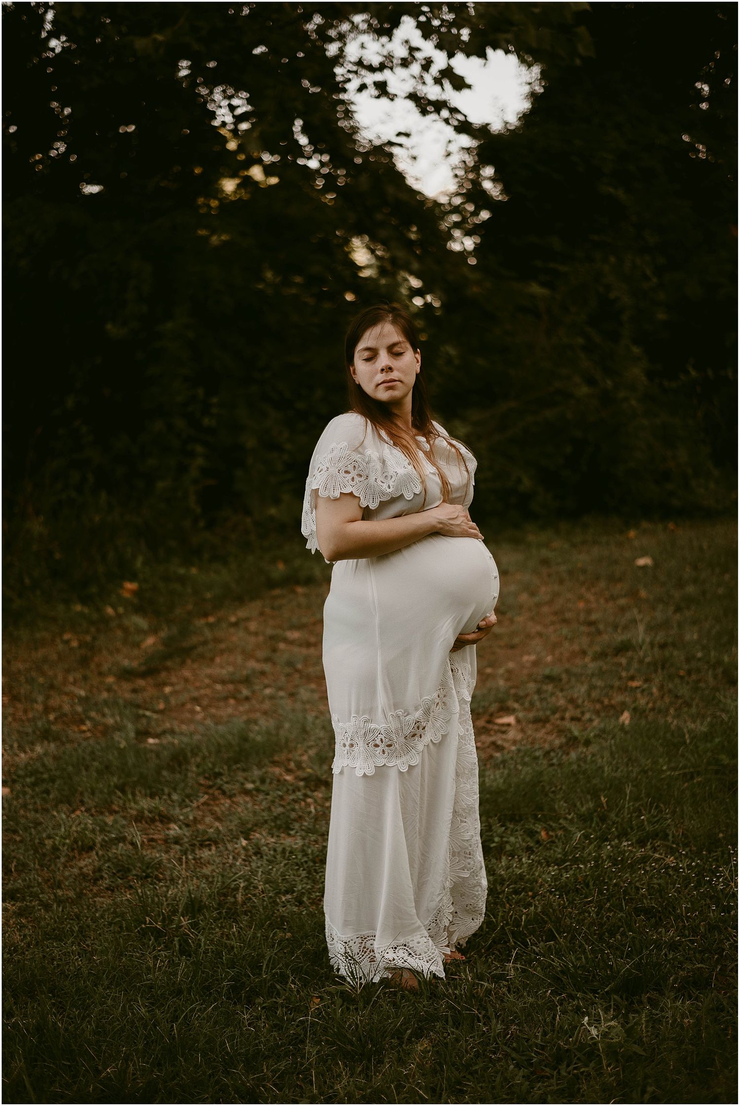 Dramatic maternity portrait with moody lighting and forest backdrop at dusk.