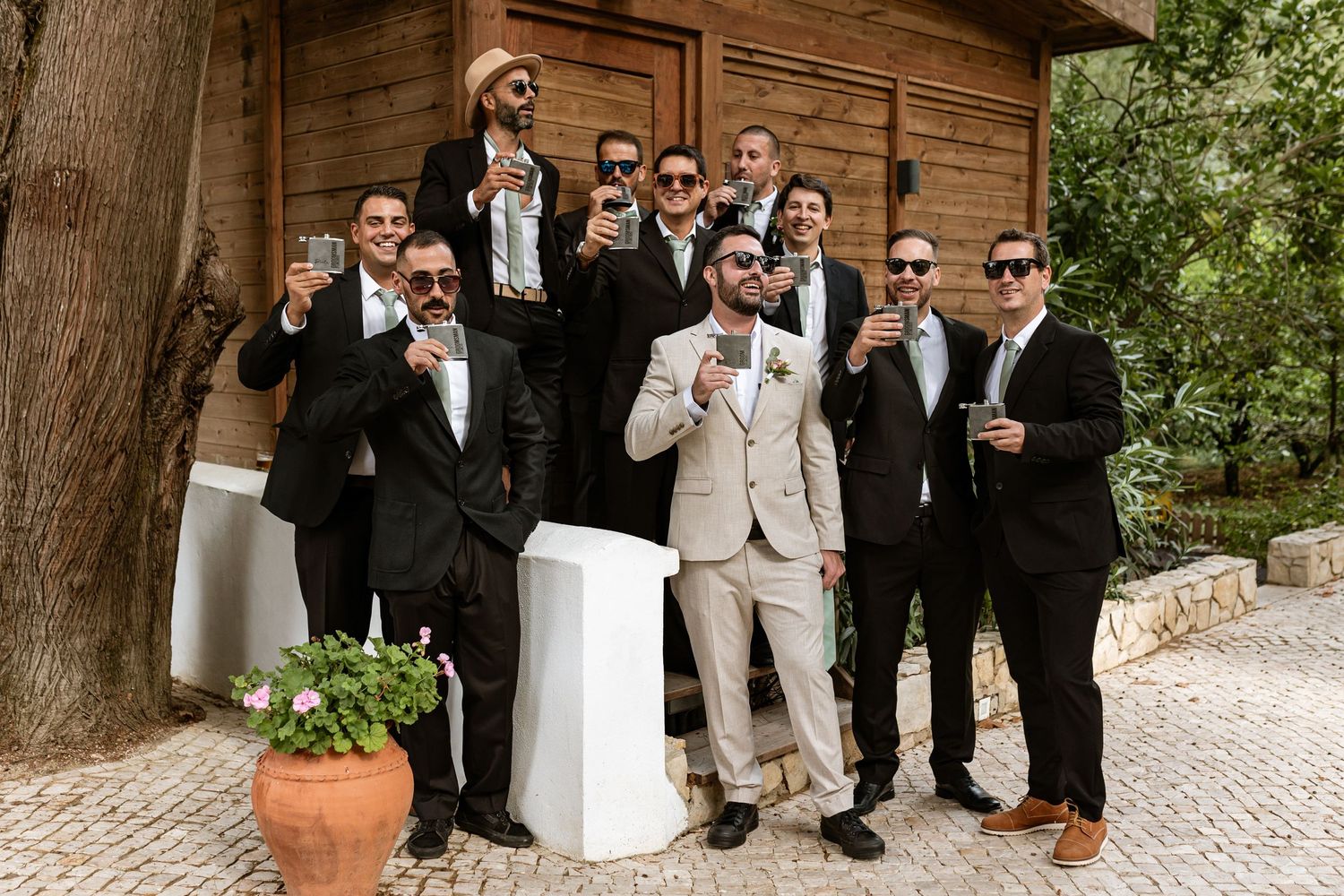 A group of well-dressed wedding party members in suits and sunglasses pose together outside a rustic wooden building at Quinta da Bichinha