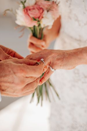 Close-up of hands exchanging wedding rings with pink and white flowers in the background.