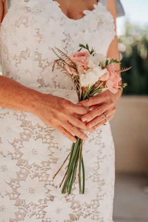 A close-up of hands holding a delicate bouquet with pink and white flowers against a lace dress.