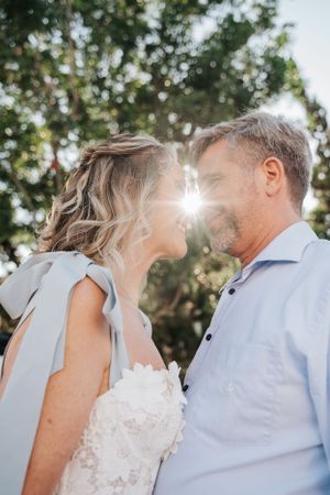 Sunlit wedding portrait with white dress and light blue shirt against garden backdrop.