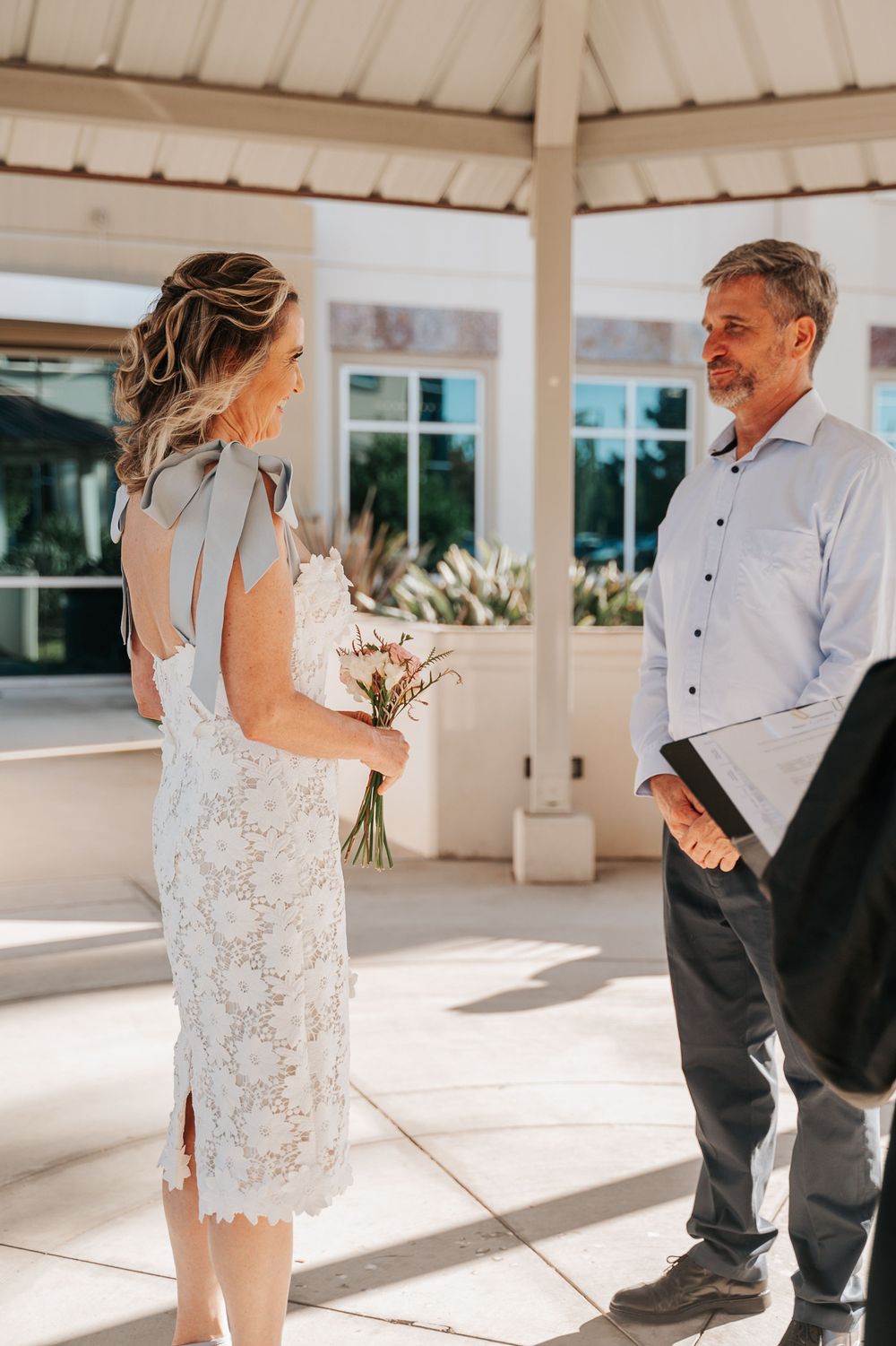 A bride in a lace dress holds a bouquet while standing under a white gazebo.