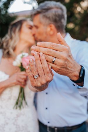 Close-up of hands displaying wedding rings with pink flowers in background.