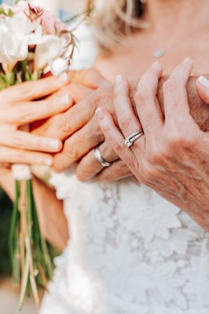 Close-up of hands with wedding rings and pink flowers against lace dress.