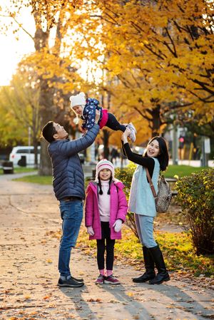An Asian family of four, mom, dad, young child standing in between them and a toddler lifted above their heads