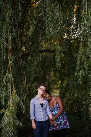 A young lesbian couple posing in front of a willow tree holding hands