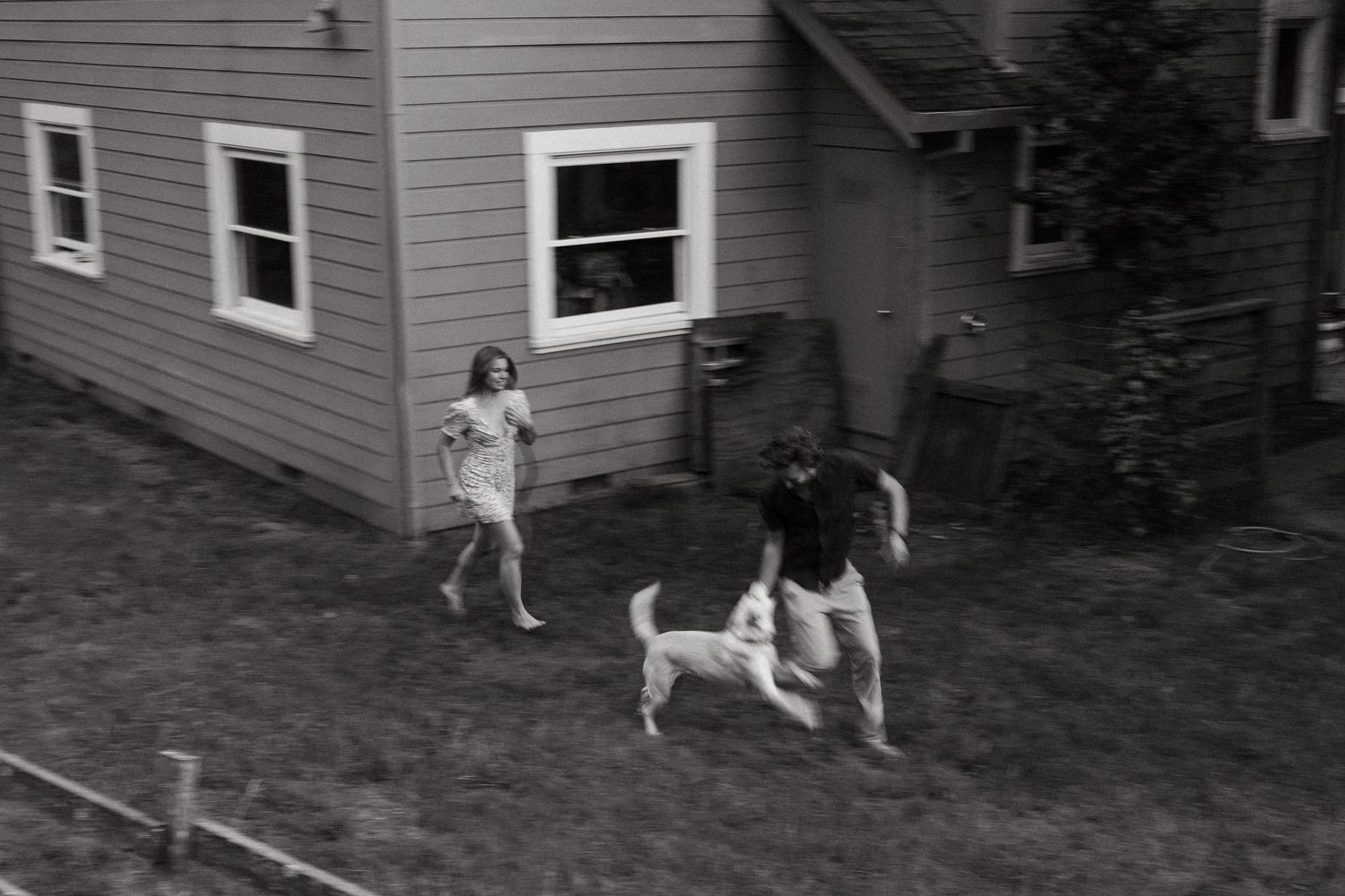 Vintage black and white photo showing people playing with a dog in a backyard near a house.