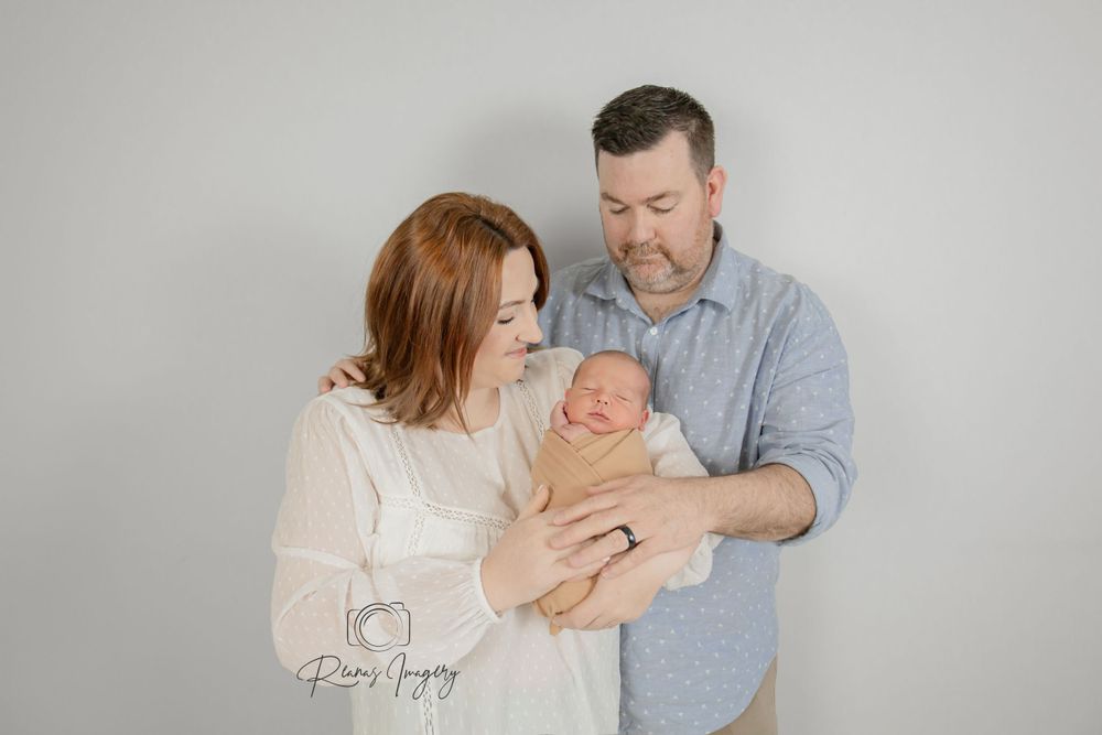 Happy new parents in white studio holding their swaddled newborn baby together, smiling lovingly.