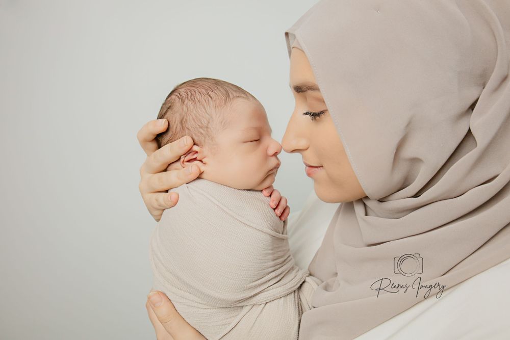 Muslim mother in beige hijab tenderly holding swaddled newborn baby close to her face in studio.