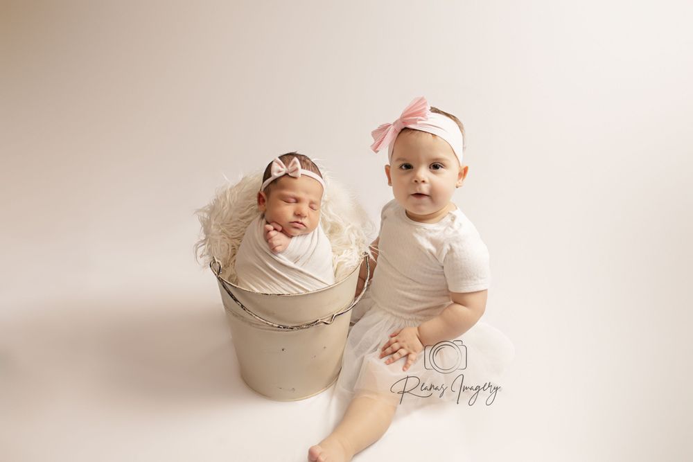 Newborn baby swaddled in white bucket beside smiling older infant sister wearing pink bow headband.