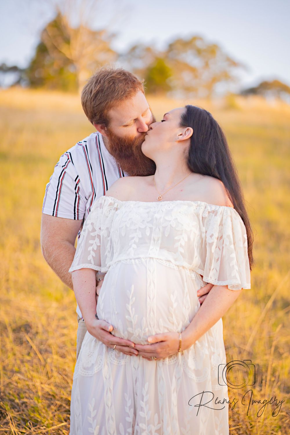 Couple kissing in golden sunlit field, woman in white off-shoulder maternity gown cradling baby bump.