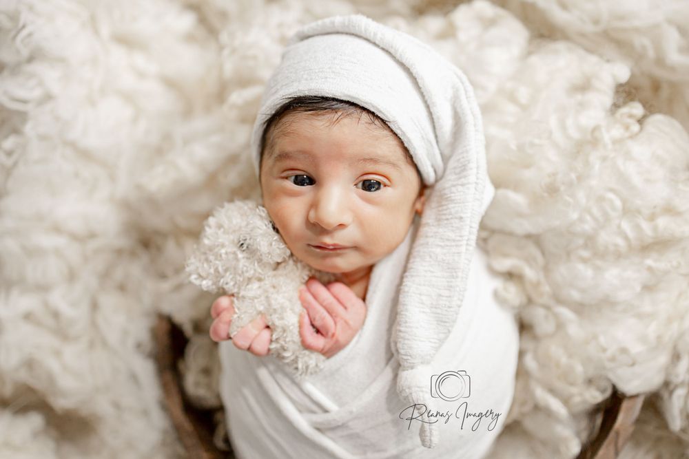 Newborn baby in white wrap and sleepy hat holds a tiny teddy bear in studio portrait.