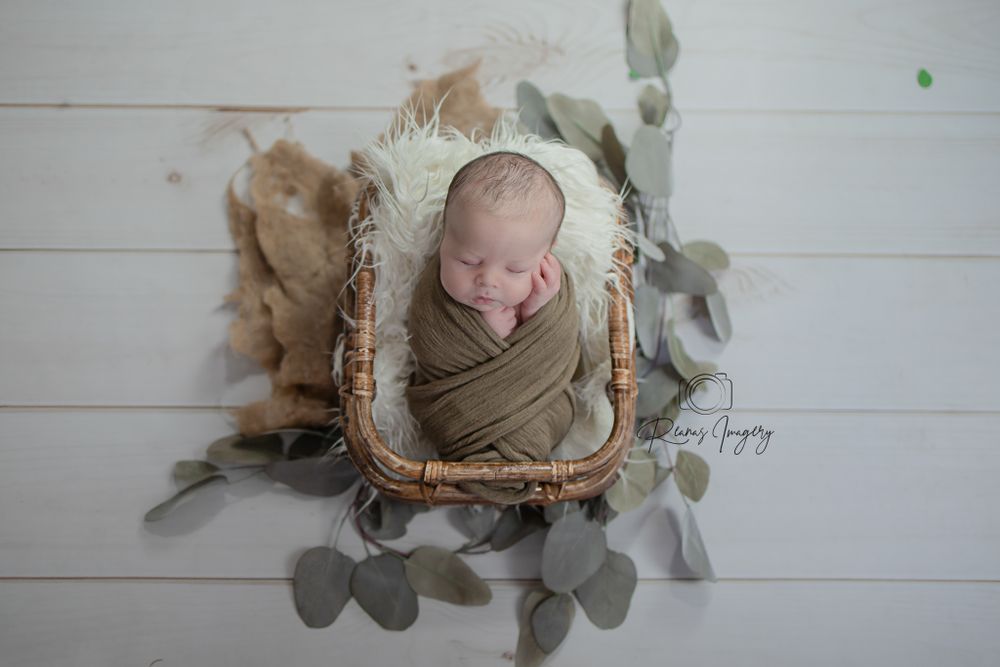 Sleeping newborn baby swaddled in olive green wrap in wicker basket with eucalyptus leaves on white wood.