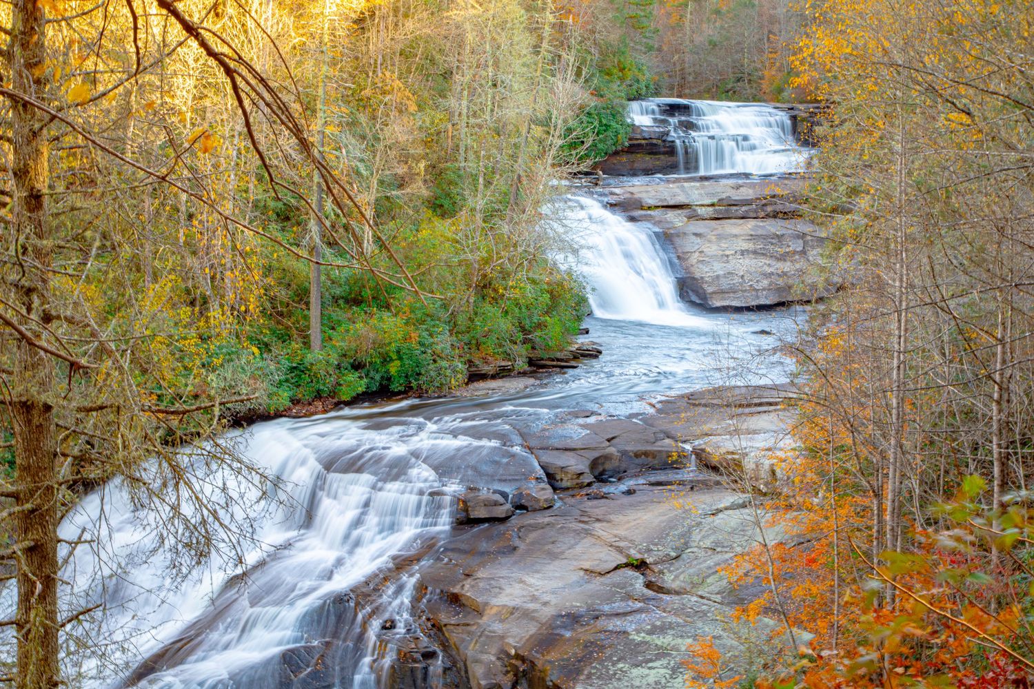 Multi-tiered waterfall flows through autumn forest with golden foliage.