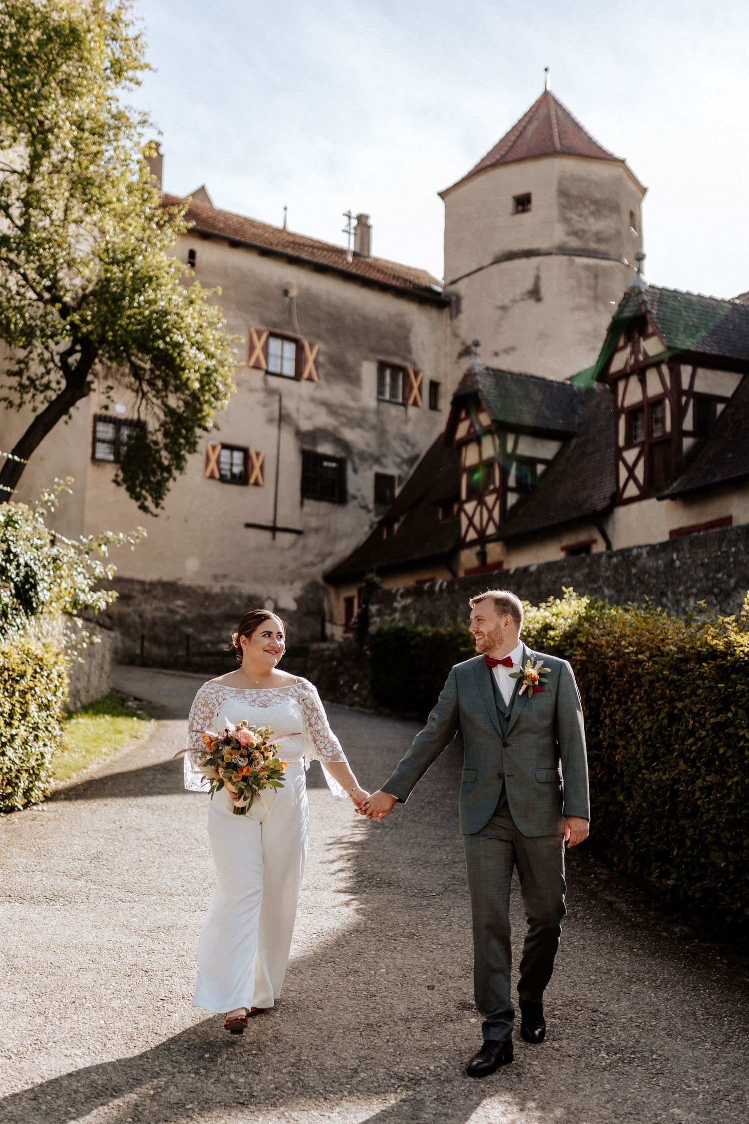 Wedding couple walks hand in hand outside historic castle with Tudor-style architecture and tower.