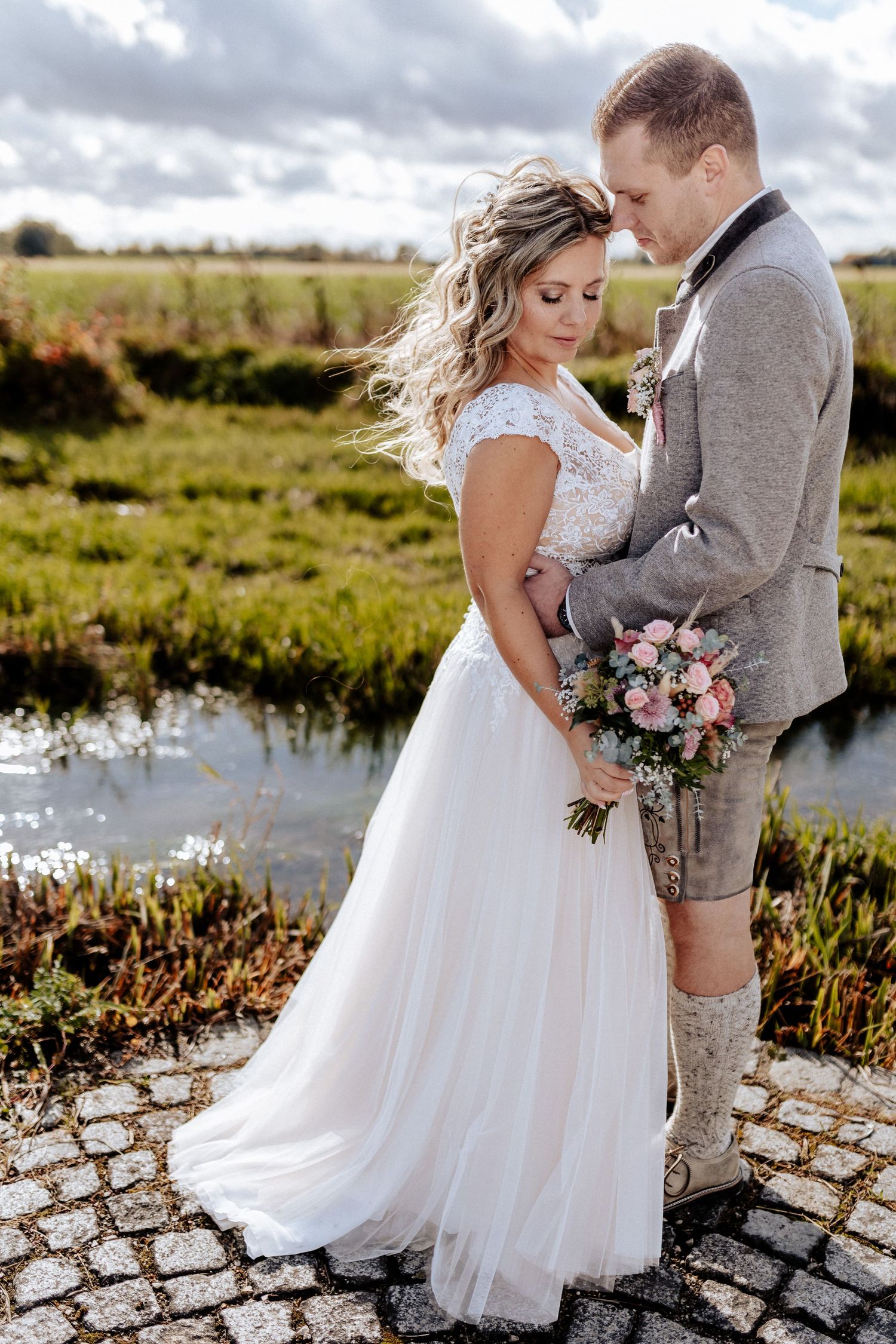 Newlyweds share an intimate moment by a pond with the bride in a flowing white dress and groom in gray attire.