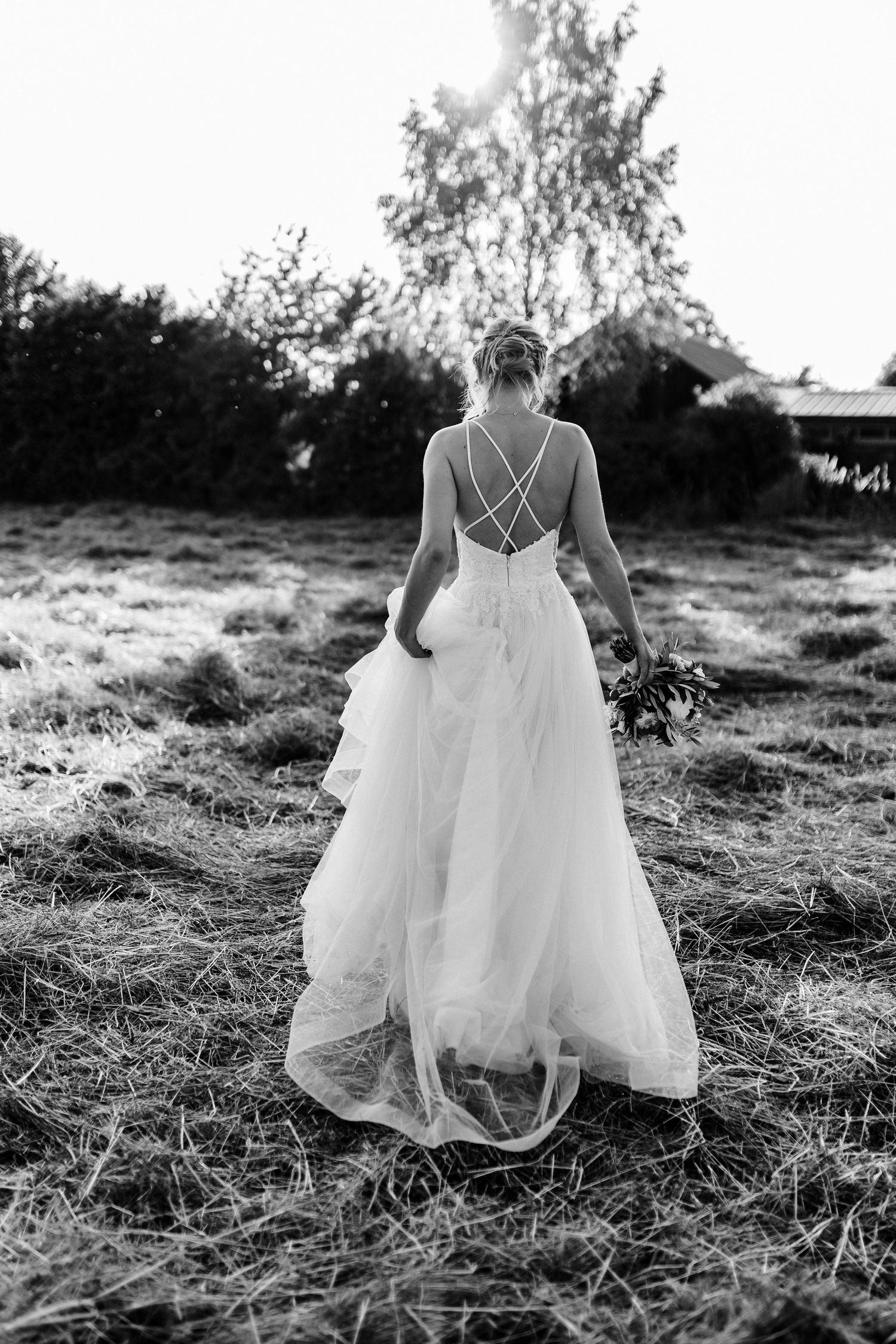 Black and white photo of bride in flowing dress walking through meadow with crossed back straps.