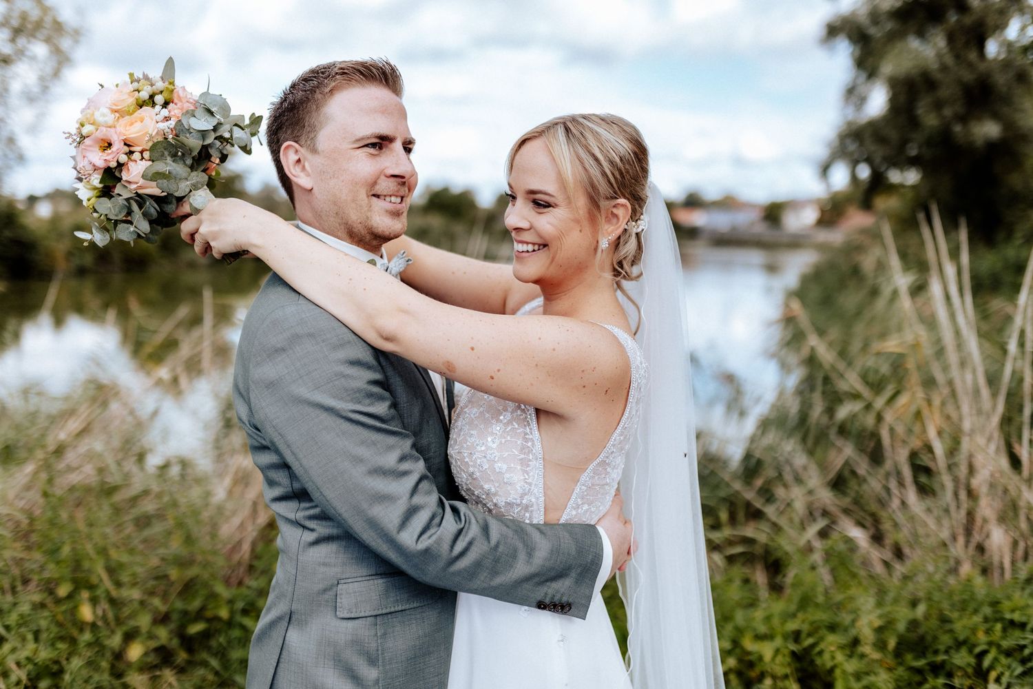 Joyful wedding couple embracing by waterside with floral bouquet.