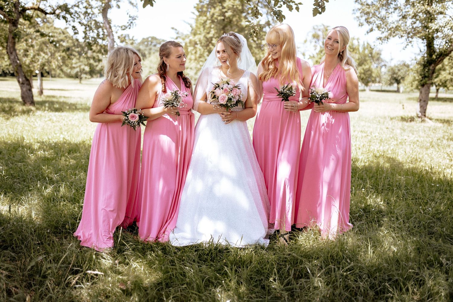 Bride in white gown poses with bridesmaids wearing matching pink dresses in sunny garden setting.