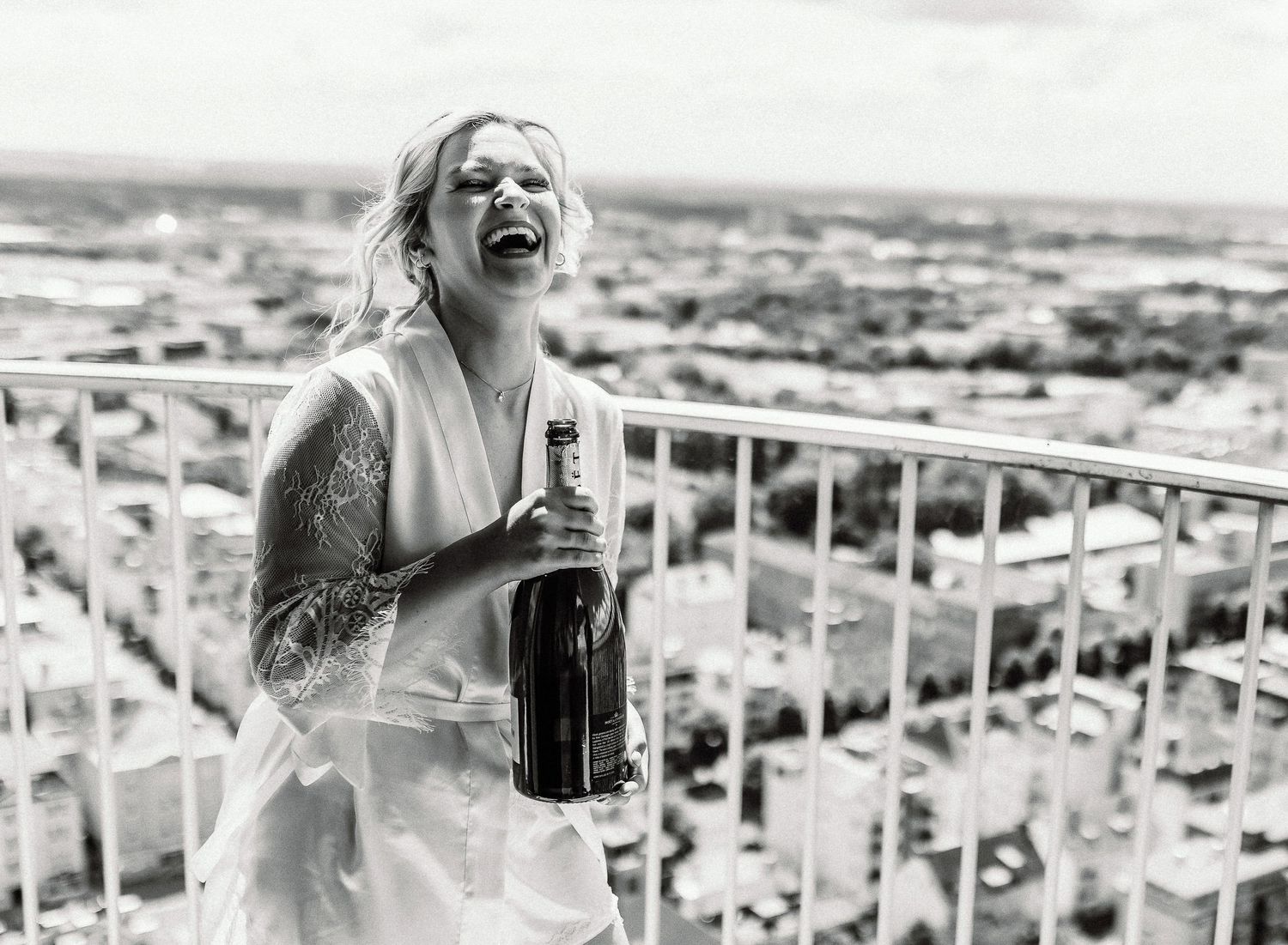 Joyful bride holding champagne bottle on balcony overlooking cityscape in black and white photograph.
