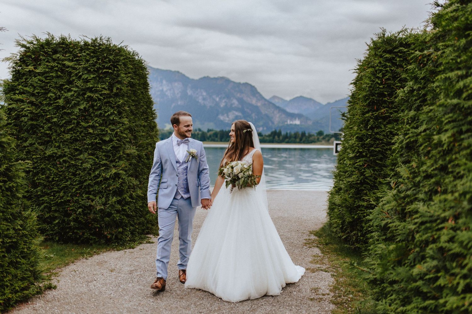 Wedding couple walks between manicured hedges with mountain lake backdrop.