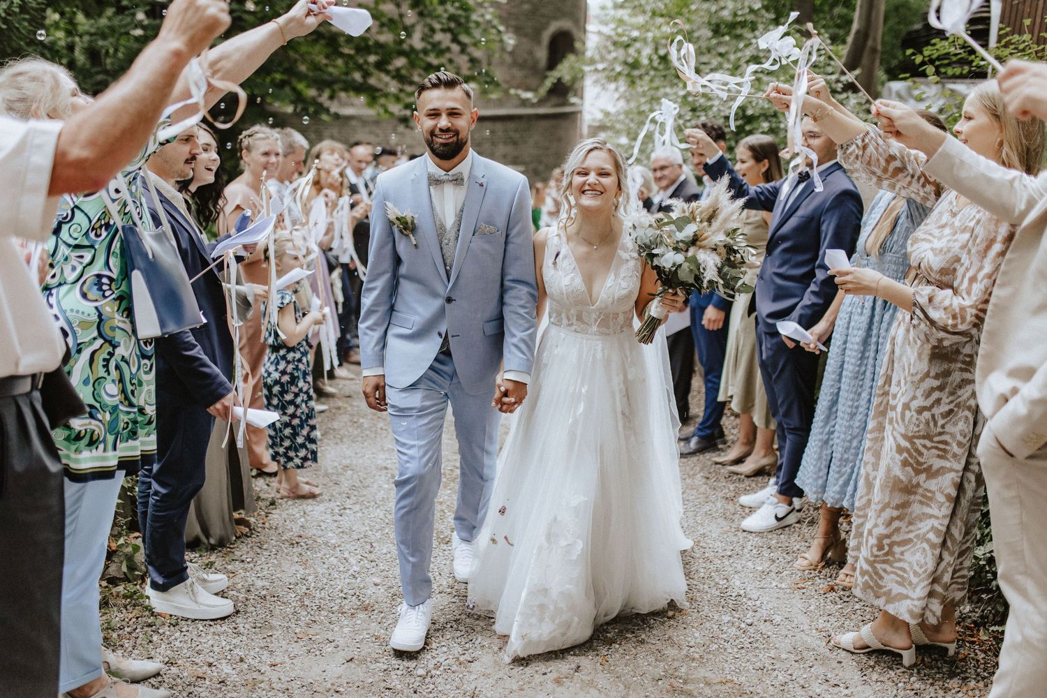 Couple walks through shower of confetti on tree-lined wedding ceremony exit path.