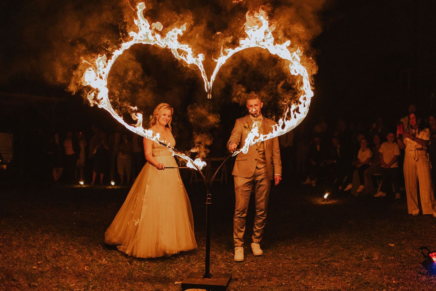 Couple creates heart-shaped light trails using fire performers at nighttime wedding celebration.