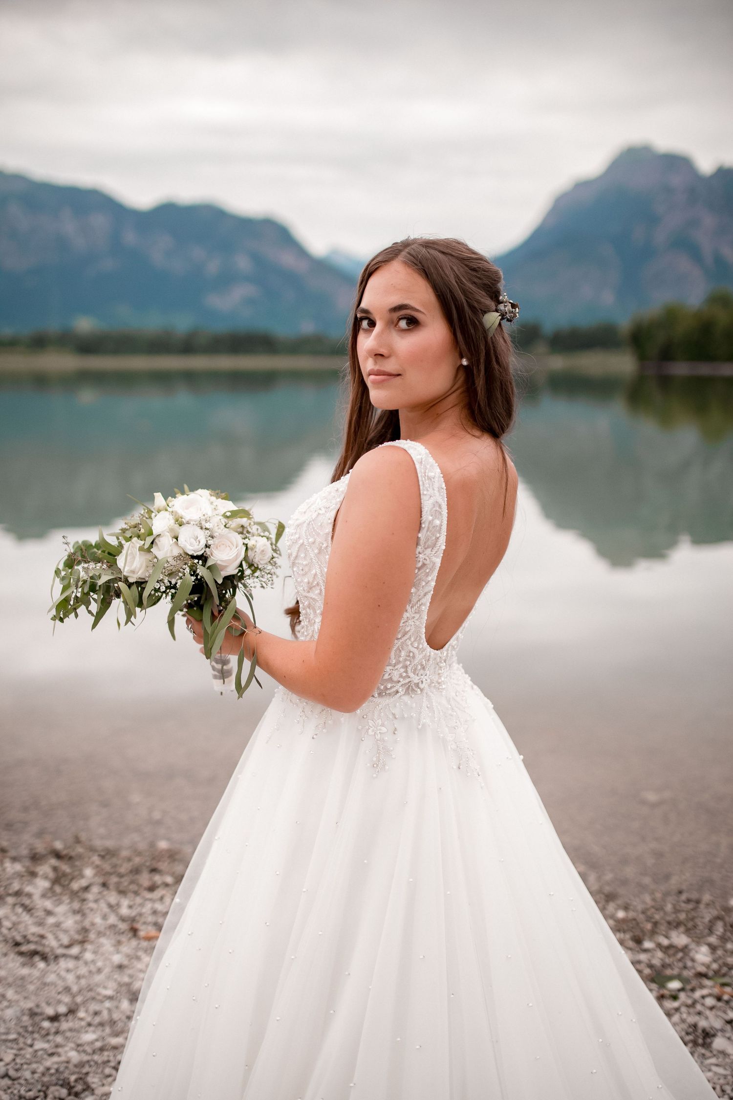 Bride in lace wedding gown poses by mountain lake with white bouquet.