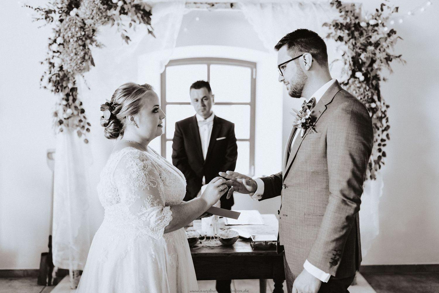 A wedding ceremony with the couple exchanging rings in front of an officiant in a bright white room.