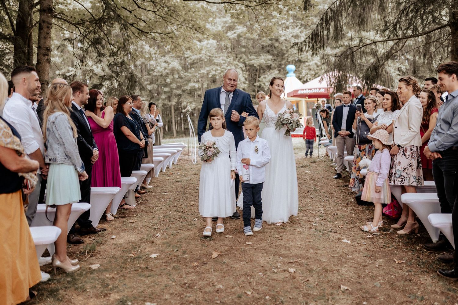 Wedding ceremony processional in a pine forest with white chairs lining the aisle.