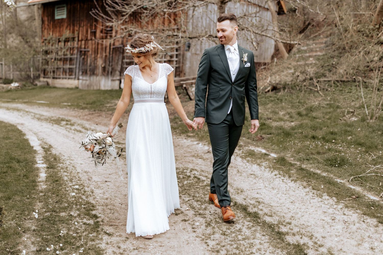 Couple walks hand in hand on rustic path in wedding attire.