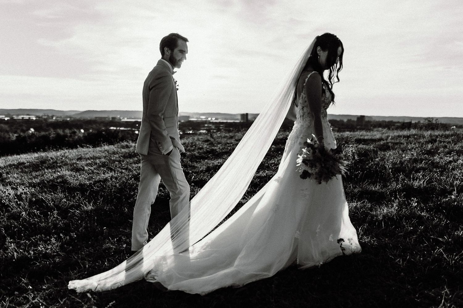 A bride and groom stand silhouetted against a scenic hilltop landscape in black and white.