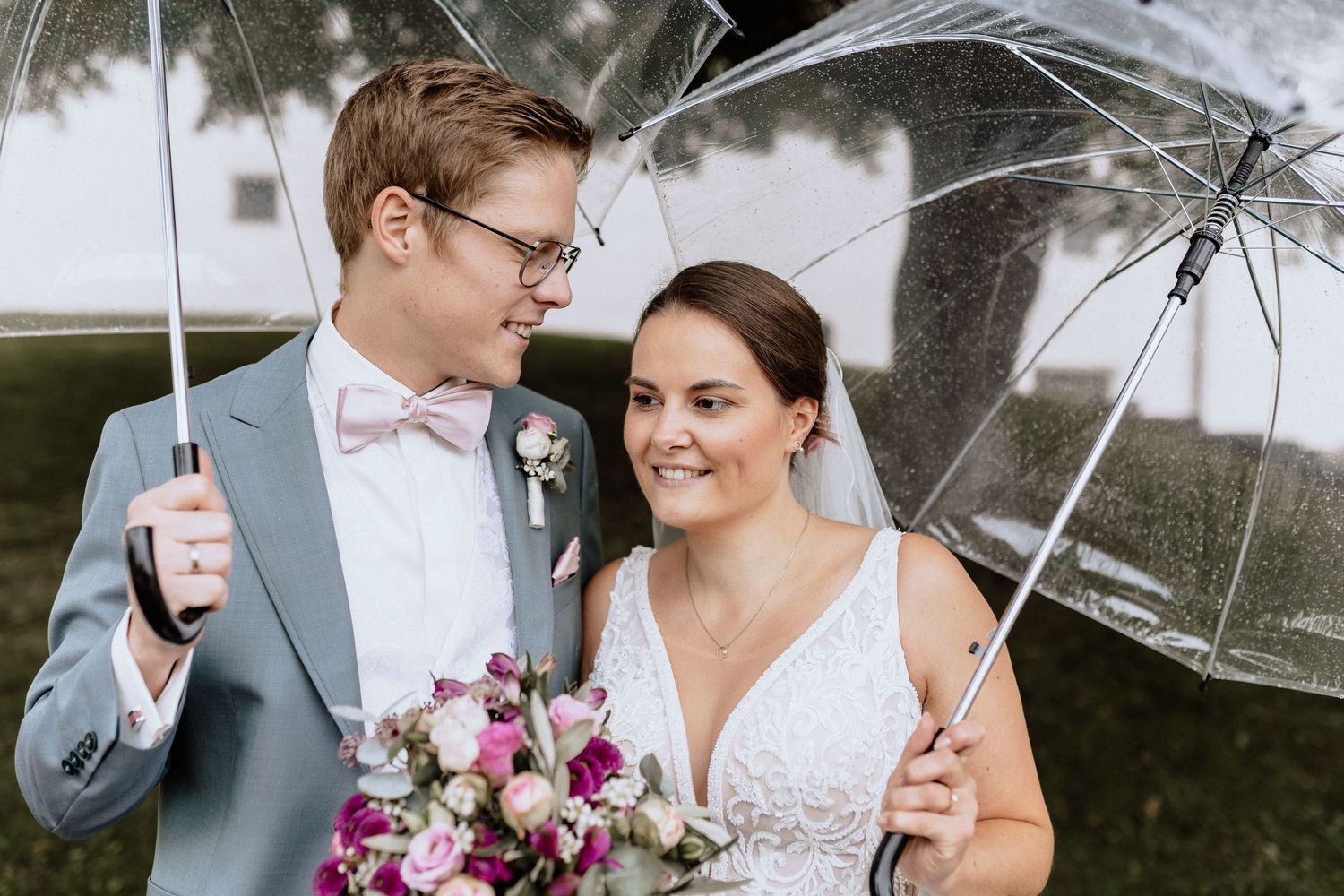 Wedding couple shares moment under clear umbrella with vibrant bouquet.