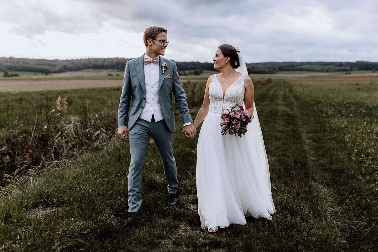 Wedding couple stands in rustic field path under moody sky.