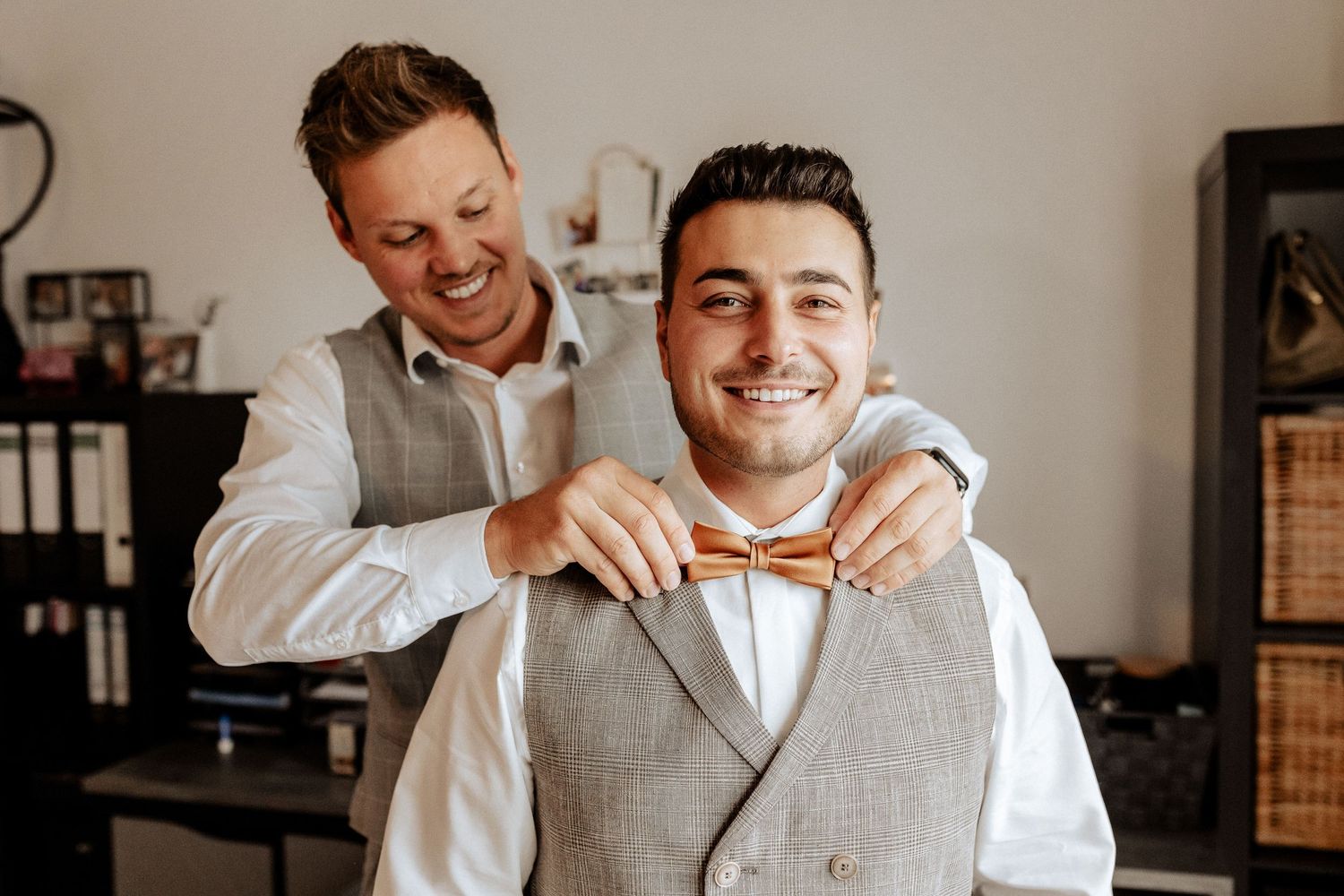 Groomsman helps adjust a bowtie during wedding preparations in a sunlit indoor setting.