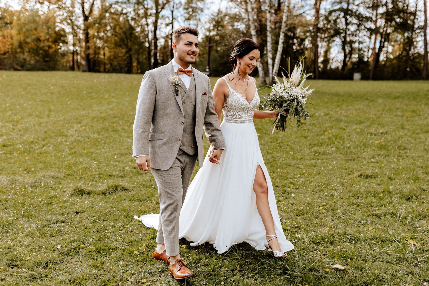 Newlywed couple walks together on grassy field with bride holding white floral bouquet.