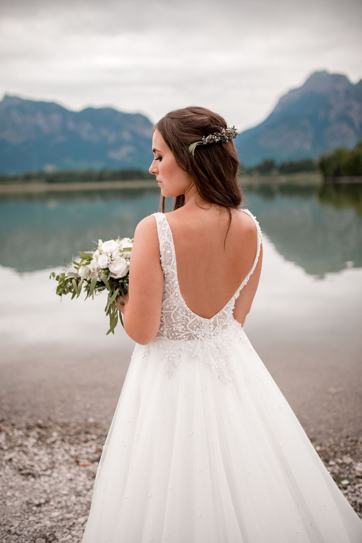 Back view of a bride in a white lace dress holding flowers against a mountain lake backdrop.