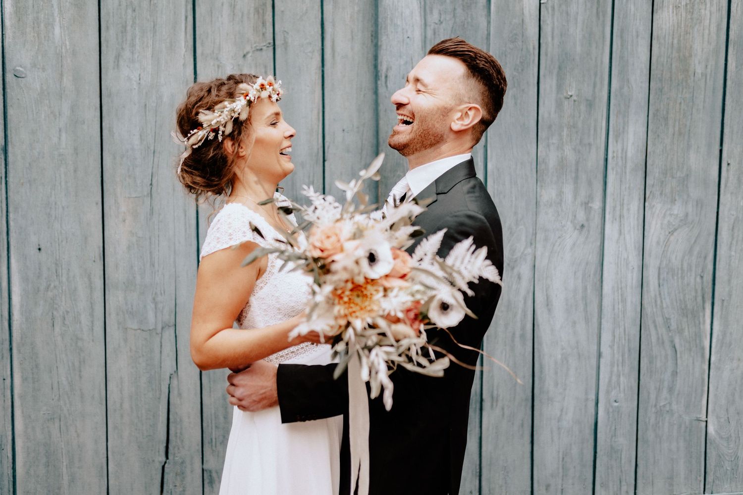 Couple sharing laughter while holding dried flower bouquet against rustic wooden wall.